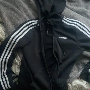 Svart huvtröja från Adidas - Säljer en svart huvtröja från Adidas med klassiska vita ränder längs ärmarna och dragkedja framtill. Tröjan har en justerbar huva och fickor, perfekt för en sportig look.