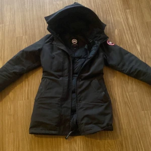 Svart dunjacka från Canada Goose - Säljer en svart dunjacka från Canada Goose med huva och dragkedja. Perfekt för kalla vinterdagar. Jackan har en ikonisk logga på ärmen och är tillverkad i Kanada.