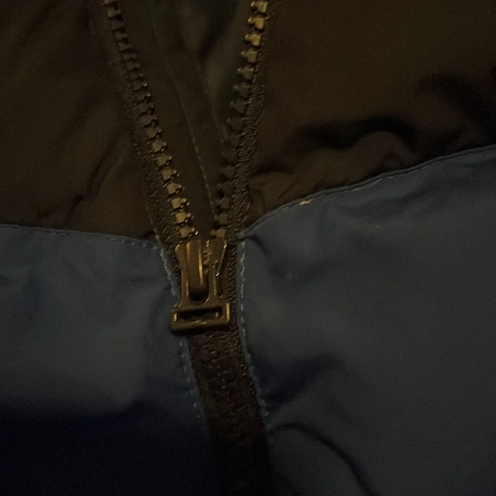 Säljer en blå och svart dunjacka från Nike med huva och dragkedja. Jackan har en sportig look och är perfekt för kyligare väder. Den har en klassisk Nike-logga på bröstet och är designad för att hålla dig varm och bekväm. Fläck på höger framsida. Takit.