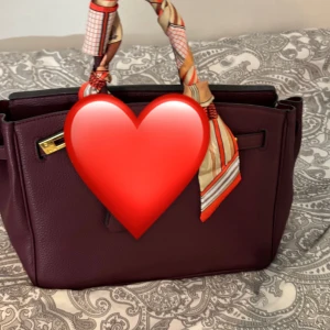 Lila handväska  - Elegant lila handväska med en färgglad scarf knuten runt handtaget. Väskan har en stilren design med guldfärgade metalldetaljer och är perfekt för att ge en färgklick till din outfit.