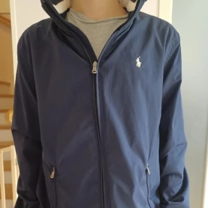 Blå jacka från Polo Ralph Lauren - Säljer en stilren blå jacka från Polo Ralph Lauren med dragkedja och logotyp på bröstet. Jackan har en hög krage och är perfekt för svala dagar. Den är långärmad och har en klassisk design. Jackan har inga defekter och är som ny.Nästan inte använd😀Perfekt vindjacka till våren.Priset är inte hugget i sten.Hör av er vid intresse👍