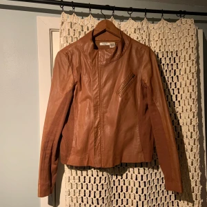 Brun skinnjacka från La Redoute - Snygg brun skinnjacka från La Redoute med dragkedja och hög krage. Jackan har en stilren design med en dragkedjeficka på bröstet och långa ärmar. Perfekt för en cool och avslappnad look.