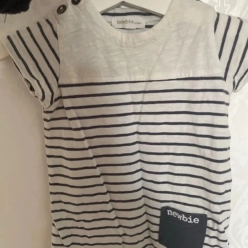 Söt randig jumpsuit från Newbie i vitt och marinblått. Jumpsuiten har korta ärmar och knappar på axlarna för en fin detalj. Den har också en liten ficka med 'newbie' tryckt på. Perfekt för en avslappnad stil.. Muu.