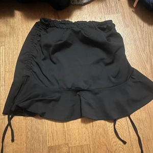 Svart kjol med volangdetaljer - Snygg svart kortkjol med volangdetaljer och knytband i sidorna. Perfekt för en stilren look.