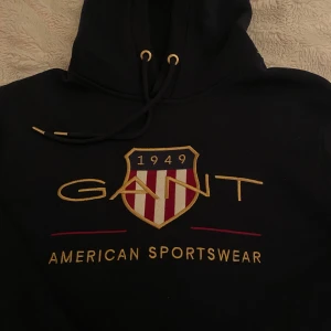 Mörkblå hoodie från GANT - Säljer en snygg mörkblå hoodie från GANT med broderad logga och texten 'American Sportswear' på framsidan. Hoodiens modell är ganska svår att hitta nu💙