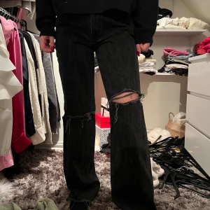 Svarta jeans med slitningar - Säljer ett par svarta jeans med slitningar på knäna. De har en bootcut-stil och är högmidjade, vilket ger en snygg och modern look. Perfekta för en avslappnad stil.