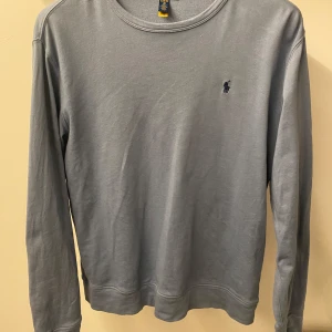 Grå tröja från Ralph Lauren - Säljer en stilren grå tröja från Ralph Lauren med det klassiska logotypbroderiet på bröstet. Tröjan har långa ärmar och en rund halsringning, perfekt för en avslappnad look. Det finns en liten fläck på magen som inte syns men därför säljer jag  den för 89 kr + den är använd mycket!
