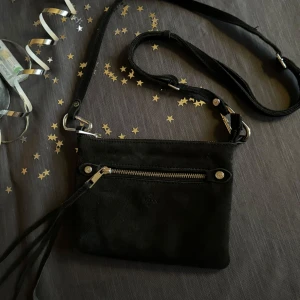 Svart axelväska från Adax - Snygg svart axelväska från Adax i mocka med justerbar axelrem. Väskan har en dragkedja upptill och en stilren design med metallknappar. Insidan är mönstrad med flera fack för enkel organisering.