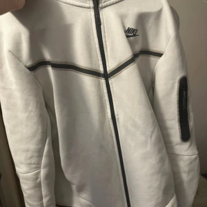Vit Nike tech hoodie - Säljer en stilren vit hoodie från Nike med dragkedja och diskret logga på bröstet. Jackan har långa ärmar och en praktisk ficka på ärmen. Perfekt för en sportig look.