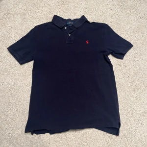 Mörkblå pikétröja från Ralph Lauren - Säljer en klassisk mörkblå pikétröja från Ralph Lauren med röd broderad logga på bröstet. Tröjan har korta ärmar och en klassisk krage med knappar. Perfekt för en stilren look. Storlek S