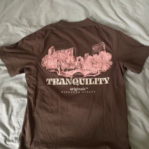 t-shirt Jack & Jones - Säljer en brun t-shirt från Jack & Jones med tryck av en stadssilhuett och texten 'Tranquility Originals Nineteen Ninety' på baksidan. T-shirten är kortärmad och gjord i bomull, perfekt för en avslappnad stil.