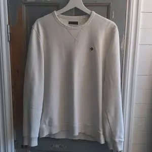 Säljer en stilren vit sweatshirt från Morris med rund hals och ett litet broderat emblem på bröstet. Tröjan har långa ärmar och ribbade muddar vid ärmslut och nederkant. Perfekt för en avslappnad look.