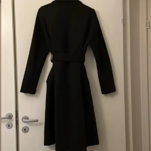 Svart kappa från Vero Moda - Snygg svart kappa från Vero Moda med ett stilrent bälte i midjan. Perfekt för att hålla stilen under kyligare dagar. Kappan har långa ärmar och en klassisk krage.