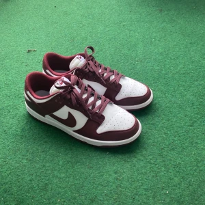 Nike Dunk i vinrött och vitt - Snygga Nike Dunk sneakers i en stilren kombination av vinrött och vitt. Skorna har klassisk snörning och en bekväm passform. Perfekta för en sportig look med en touch av stil.