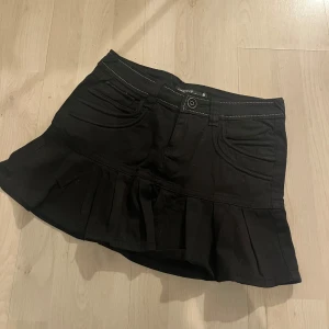 Svart kortkjol med volang - Snygg svart kortkjol med volangdetaljer. Kjolen har fickor fram och bak samt en knapp och dragkedja. Midjemått 38, längd 34cm storlek 27 