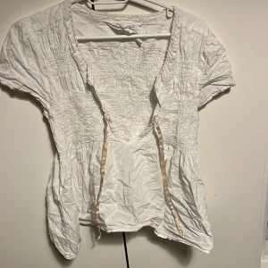 Vit blus från Odd Molly - Säljer en söt vit blus från Odd Molly med korta ärmar och knytband framtill. Blusen har en smockdetalj över bröstet och är tillverkad i ett lätt och luftigt material. Perfekt för en avslappnad och stilren look.