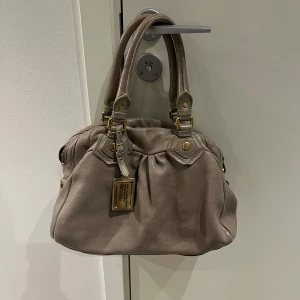 Beige handväska med gulddetaljer - Säljer en snygg beige handväska från Marc by Marc Jacobs. Väskan har gulddetaljer och en praktisk dragkedja. Insidan är fodrad med ett mönster av bokstäver. Perfekt för att bära dina dagliga nödvändigheter med stil. På guldplattan saknas det en skruv men ingenting man tänker på. Väskan är även  smutsig på insidan🩷