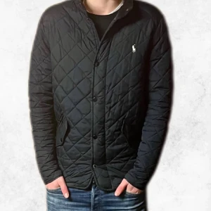 Svart quiltad jacka från Ralph Lauren - Säljer en marinblå/svart quiltad jacka från Ralph Lauren. Den är perfekt nu till våren. Den är i bra skick förutom ett litet hål i vänster ficka som inte syns från utsidan. Jackan har en liten skada på ärmen som jag har försökt sy ihop. Resultatet är som på sista bilden. Personen på bilden är 185 cm så den passar någon som är runt den längden.