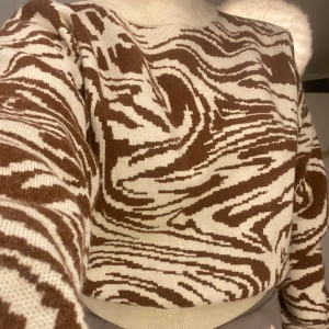 Brun och vit mönstrad tröja - Säljer en snygg tröja med ett unikt brunt och vitt mönster. Tröjan har en hög krage och långa ärmar, perfekt för kyligare dagar. Den är både stilren och bekväm, vilket gör den till ett måste i garderoben.