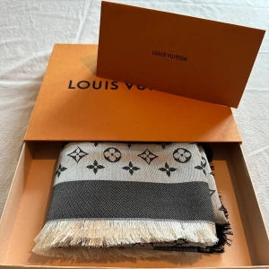 Grå och svart sjal från Louis Vuitton - Elegant grå och svart halsduk från Louis Vuitton med det ikoniska monogrammönstret. Tillverkad i en lyxig blandning av siden och ull, vilket ger en mjuk och varm känsla. Perfekt för att ge en touch av lyx till din outfit.