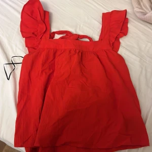 Röd topp med volangdetaljer från H&M - Säljer en röd topp från H&M med volangdetaljer på axelbanden och knytning i ryggen. Toppen har en luftig passform och är perfekt för varma dagar. Materialet är lätt och bekvämt.