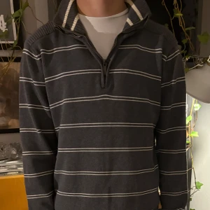 Grå randig tröja med dragkedja - Säljer en stilren grå tröja med vita ränder och dragkedja vid halsen. Tröjan har en hög krage och långa ärmar, perfekt för kyligare dagar. Den ribbade kragen ger en extra touch av stil. Passar bra till både jeans och chinos.
