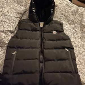 Svart dunväst från Moncler - Snygg svart dunväst från Moncler med dragkedja och huva. Västen har Moncler-logga på bröstet och text på insidan av huvan. Perfekt för kyligare dagar med stil.