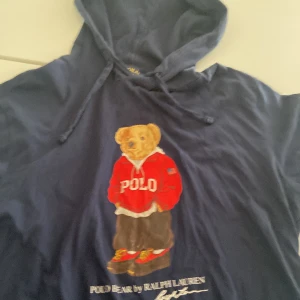 Blå hoodie med björn från Ralph Lauren - Säljer en blå hoodie från Ralph Lauren med ett charmigt björnmotiv på framsidan. Tröjan har långa ärmar och en klassisk huva med dragsko. Perfekt för en avslappnad stil.