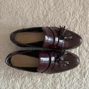 Bruna loafers med tofsar från Zara - Snygga vinröda loafers från Zara med dekorativa tofsar på ovansidan. Skorna har en glansig finish och en robust sula som ger bra stöd. Perfekta för en stilren look.