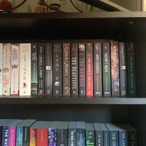 Bokhylla med fantasy och ungdomslitteratur - En bokhylla fylld med fantasy och ungdomsböcker. Titlar som 'Crave', 'The Beautiful' och 'And I Darken' syns tydligt. Böckerna har färgglada och mörka omslag, vilket ger en mystisk och lockande känsla. Perfekt för den som älskar spänning och magi. Priset varierar beroende på vilken bok man vill ha💕