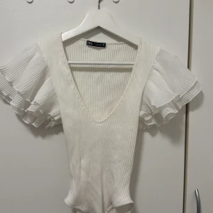 Vit ribbad body från Zara - Säljer en stilren vit ribbad body från Zara med v-ringning och korta volangärmar. 