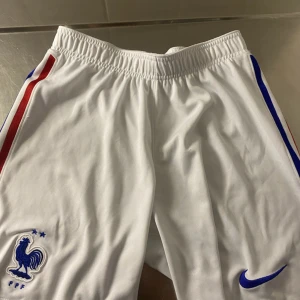 Vita fotbollsshorts från Nike - Snygga vita fotbollsshorts från Nike med Dri-FIT-teknologi. Shortsen har blå och röda ränder längs sidorna och en broderad Nike-logga. Perfekta för fotbollsträningen eller matchen. 🇫🇷
