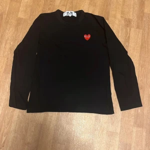 Comme des Garçons långärmad tröja - Säljer en svart långärmad tröja från Comme des Garçons med ett ikoniskt rött hjärta broderat på bröstet. Perfekt för en stilren och trendig look. Passar bra till jeans