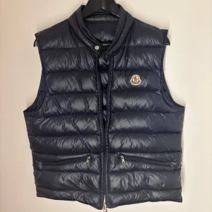 Blå dunväst från Moncler - Säljer en snygg blå dunväst från Moncler. Västen har en quiltad design med dragkedja och två praktiska fickor framtill. Perfekt för kyligare dagar när du vill hålla stilen. OBS dragkedjan funkar ej går att fixa billigt och enkelt men bara inte årkat därav priset. Priset är diskuterbart!