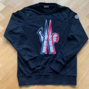 Svart sweatshirt från Moncler - Säljer en svart sweatshirt från Moncler med ett stort broderat märke i rött, vitt och blått på framsidan. Tröjan har långa ärmar och en rund halsringning. Även en logga på axeln. Storlek M. Priset kan diskuteras. Knappast använd, inga tecken på slitningar eller skador.