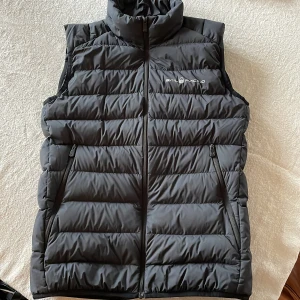 Svart dunväst från Sail Racing - Snygg svart dunväst från Sail Racing med dragkedja och hög krage. Västen har två praktiska fickor med dragkedja och är perfekt för kyligare dagar. Tillverkad med högkvalitativt dun från Allied Feather & Down.