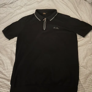 Svart pikétröja från Loro Piana - Säljer en stilren svart pikétröja från Loro Piana. Den har en klassisk krage med dragkedja och korta ärmar. Perfekt för både vardag och lite finare tillfällen. Tröjan är i bra skick och passar till det mesta i garderoben. Skicka ett meddelande vid intresse!