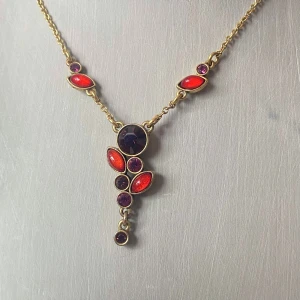 Guldhalsband med röda och lila stenar - Snyggt guldhalsband med en unik design av röda och lila stenar. Perfekt för att ge en färgklick till din outfit. Kedjan är tunn och elegant, vilket gör det lätt att bära både till vardags och fest. Stenarna är arrangerade i en vacker formation som ger halsbandet en exklusiv look.