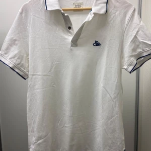 Vit pikétröja från edc - Säljer en stilren vit pikétröja från edc. Den har korta ärmar och en klassisk krage med blå detaljer. Tröjan är i bomull och har en broderad logga på bröstet. Perfekt för sommardagar eller en avslappnad vårlook. Passar både till jeans och shorts!