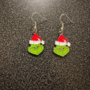 Grinch örhängen med tomteluva - Säljer ett par roliga örhängen med Grinch-motiv. De är gröna med en röd och vit tomteluva, perfekta för jul och nyårsfester. Örhängena har en krok för enkel användning. Perfekt för att sprida lite humor under högtiderna! 🎄Krokarna är inköpta som nickelsäkra.