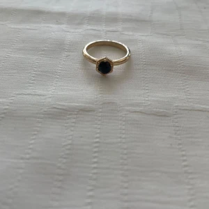 Guldfärgad ring med svart sten - Säljer en elegant guldfärgad ring med en stor svart sten i mitten. Ringen har en klassisk och stilren design som passar perfekt för både vardag och fest. Den svarta stenen är fasetterad och omgiven av en fin kant som ger extra glans. Perfekt accessoar för att lyfta din outfit!