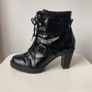 Svarta boots med klack och snörning - Snygga svarta boots  med klack och snörning. De har en cool spänndetalj och dragkedja på sidan för enkel på- och avtagning. Perfekta för höst och vinter, både till vardags och fest. De är i bra skick och redo för nya äventyr!