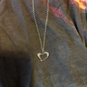 Silverfärgat halsband med hjärthänge - Säljer ett superfint silverfärgat halsband med ett hjärthänge. Hänget är dekorerat med små glittrande stenar som ger en elegant touch. Perfekt för både vardag och fest! Kedjan är tunn och smidig, vilket gör den bekväm att bära hela dagen. Passar perfekt som present eller för att skämma bort dig själv. 💖