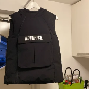 Svart väst från Hoodrich - Säljer en svart väst från Hoodrich i storlek L för åldrarna 12-13 år. Västen har en stor ficka framtill med Hoodrich-loggan och en huva. Perfekt för en cool och avslappnad stil. Använd den över en hoodie för extra värme och stilpoäng!