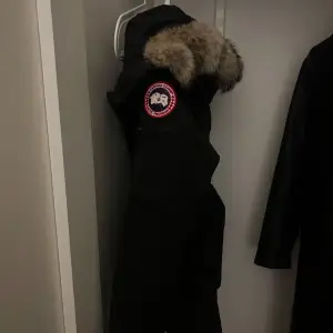 Säljer en svart parkajacka från Canada Goose med pälsdetaljer på huvan. Jackan har en klassisk design med dragkedja och knappar framtill. Perfekt för kalla vinterdagar. Håll dig varm och stilren! 🖤