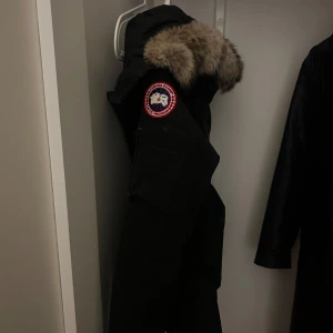 Svart parkajacka med päls från Canada Goose - Säljer en svart parkajacka från Canada Goose med pälsdetaljer på huvan. Jackan har en klassisk design med dragkedja och knappar framtill. Perfekt för kalla vinterdagar. Håll dig varm och stilren! 🖤