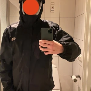 Svart vindjacka från The North Face - Säljer en svart vindjacka från The North Face i mycket bra skick. Jackan har en praktisk huva och dragkedja framtill. Perfekt för höst och vår, med en stilren design som passar till det mesta. Den är långärmad och gjord i ett slitstarkt syntetmaterial. Tål även vatten. Dragkedjan kan strula men inga problem så länge man sätter in den på rätt sätt