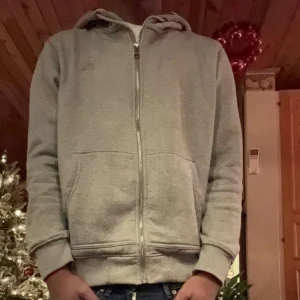 Grå Burberry Hoodie - Säljer en stilren grå hoodie från Burberry i storlek M. Den har en dragkedja framtill och en klassisk rutig insida i huvan. Perfekt för både vardag och lite kyligare dagar. Passar bra till jeans eller joggers. Hör av dig vid intresse!
