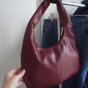 Vinröd axelväska i syntetmaterial - Snygg vinröd axelväska med ett coolt quiltat mönster. Perfekt för höst och vinter, och den har en praktisk dragkedja. Väskan är i syntetmaterial och har en modern stil som passar både till vardags och fest. Köptes för 600, säljer för 350 