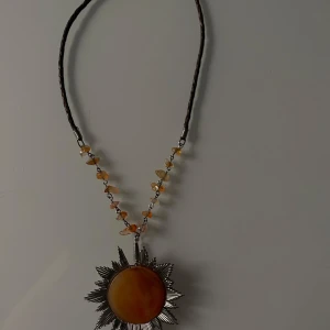 Halsband med solformad hänge - Unikt halsband med ett stort solformad hänge i mitten. Hänget har en varm gul färg och är omgivet av strålande silverdetaljer. Kedjan är dekorerad med små pärlor i orange och silver, vilket ger en bohemisk känsla. Perfekt för att liva upp en enkel outfit eller som ett statement piece på festen.